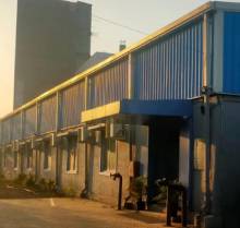 A ground-level view of a long shed with grey walls, a blue and silver corrugated metal roof, and multiple windows and entryways. The shed is situated on a paved area, and a taller, darker building is visible in the hazy background under a light sky.