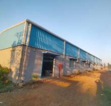 	A wide, ground-level shot of a long, light-colored shed with a blue corrugated metal roof under a clear blue sky. The shed has several small windows along its side and a large open doorway. The surrounding ground is uneven and appears to be dirt and sparse vegetation.
