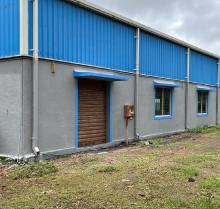 	A close-up view of the side of an shed featuring a blue corrugated metal roof and grey walls. There is a closed brown roller shutter door with a blue frame, two small windows with blue frames, and some electrical fixtures on the exterior wall. The ground in front of the building is covered with patchy green grass. The sky is overcast.