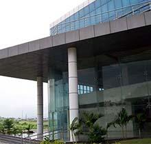 A close-up view of the modern entrance of a multi-story building, likely the IP Tower in Talawade, Pune, showcasing a flat, dark overhang supported by large, white cylindrical pillars. Expansive glass windows reflect the interior and some exterior elements. Small palm trees and other greenery are planted in front of the entrance. The upper portion of the building features reflective blue glass. In the background, a glimpse of the surrounding landscape with some vegetation and possibly a road is visible under a bright sky.