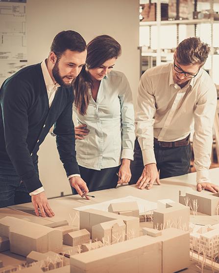 Three individuals are gathered around a table, closely examining an architectural model.The model features detailed buildings, structures, and miniature landscaping elements, such as trees. The scene suggests a collaborative discussion about design or planning.