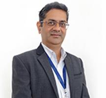 a person standing against a white backdrop, dressed in a dark grey blazer and white shirt, with a blue lanyard around their neck.