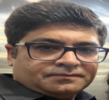 a person with dark hair is seated on a red and white patterned chair. Behind them, there is a decorative element, possibly a flower arrangement, suggesting a formal or elegant setting.